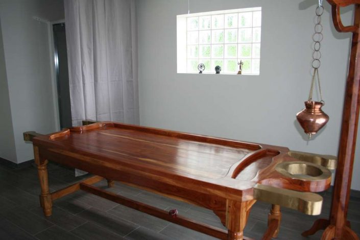 Droni (table traditionnelle pour les massages ayurvédiques) et l’espace douche en arrière plan